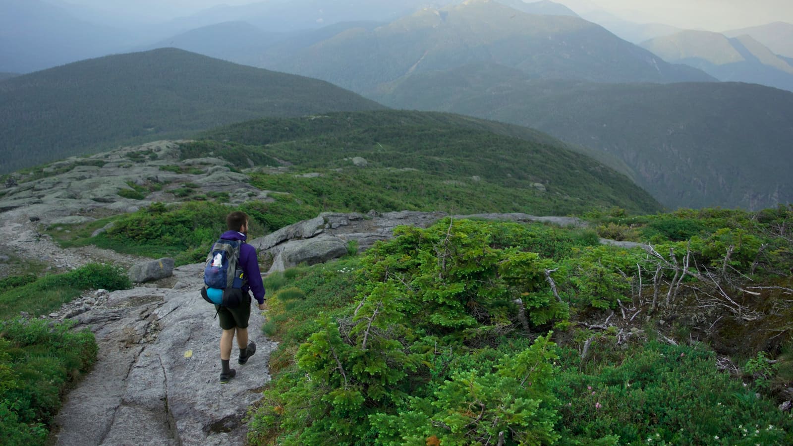 A hiker with a blue pack walking a rocky ridge trail through green mountains under a moody sky