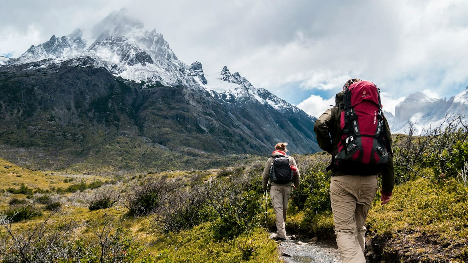 A backpacker with a large red pack hiking a trail toward a snow-capped peak