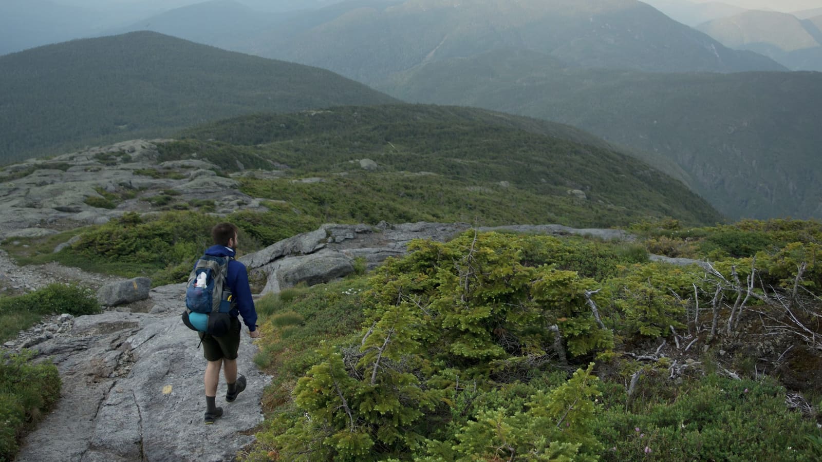 A hiker with a blue pack walking a rocky ridge trail through green mountains under a moody sky
