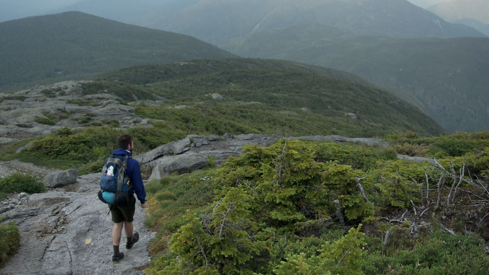 A hiker with a daypack climbing a rocky alpine trail above treeline in green mountains