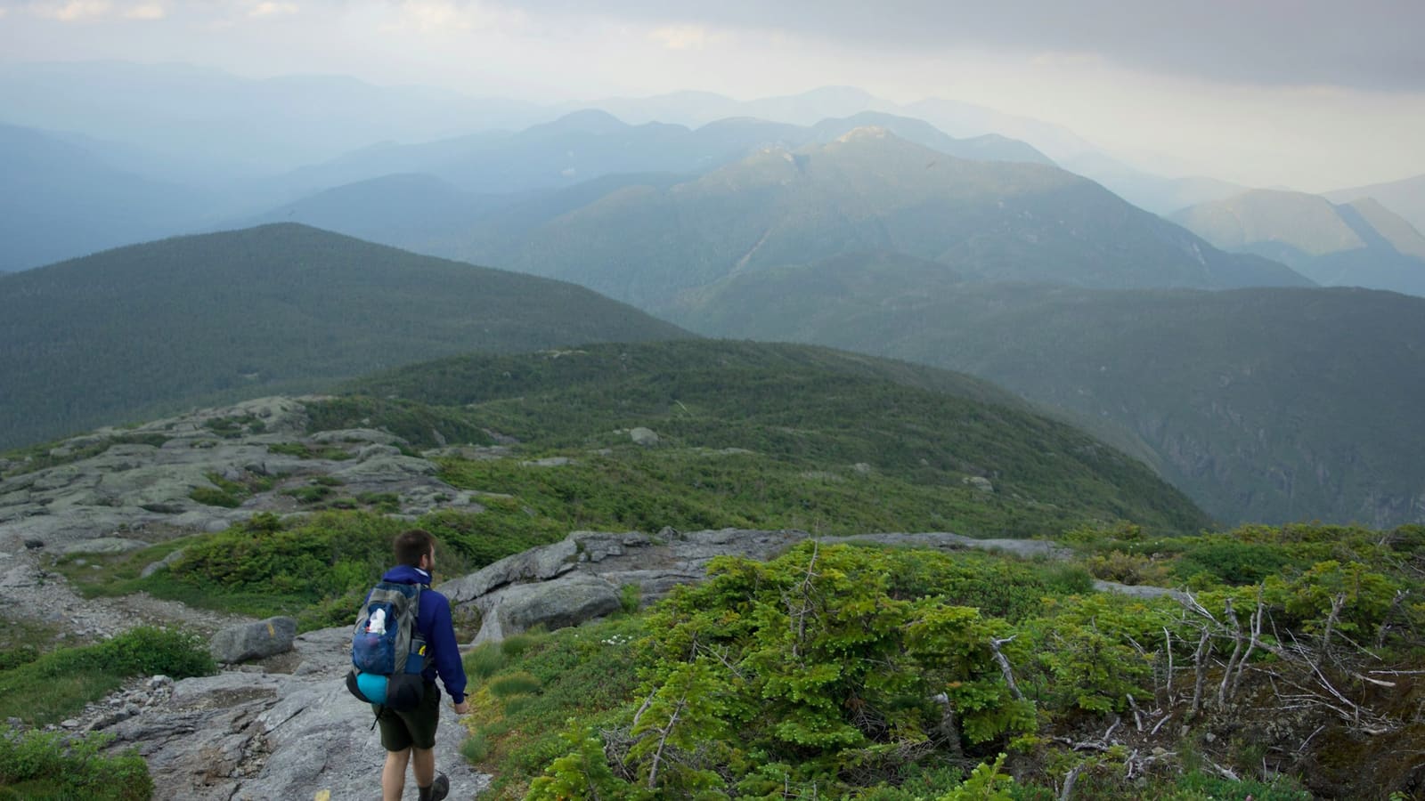 A hiker with a blue pack walking a rocky ridge trail through green mountains under a moody sky