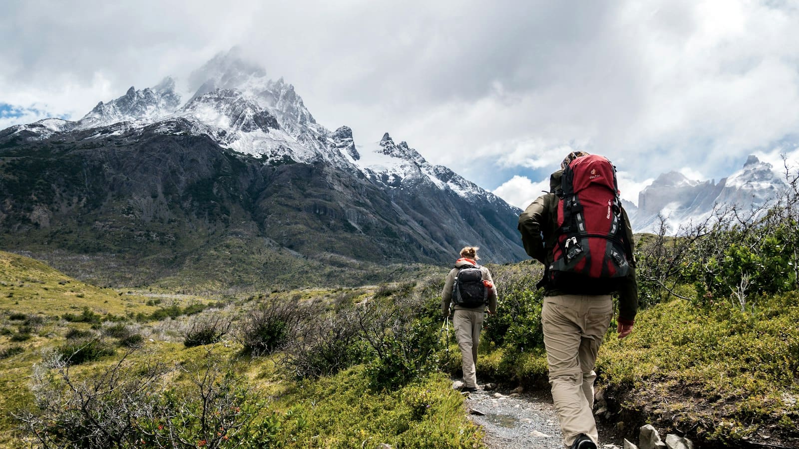 A backpacker with a large red pack hiking a trail toward a snow-capped peak