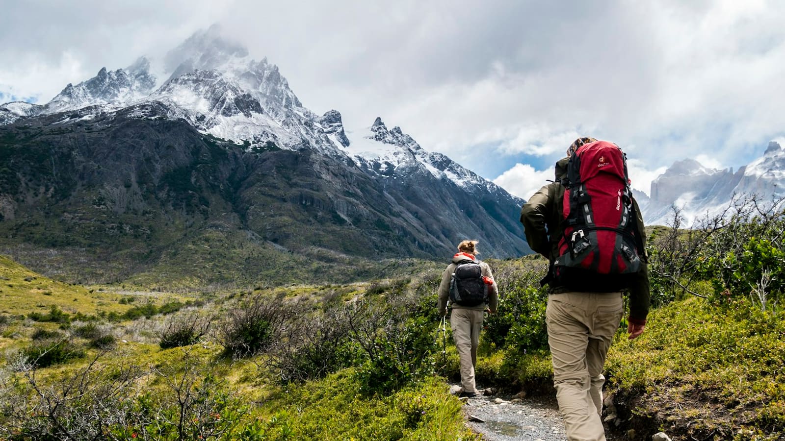 A backpacker with a large red pack hiking a trail toward a snow-capped peak