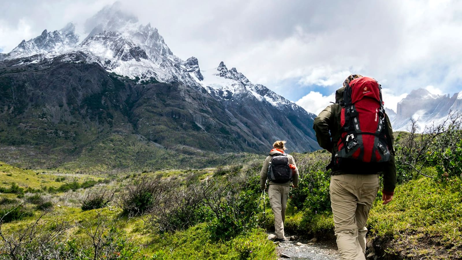 A backpacker with a large red pack hiking a trail toward a snow-capped peak