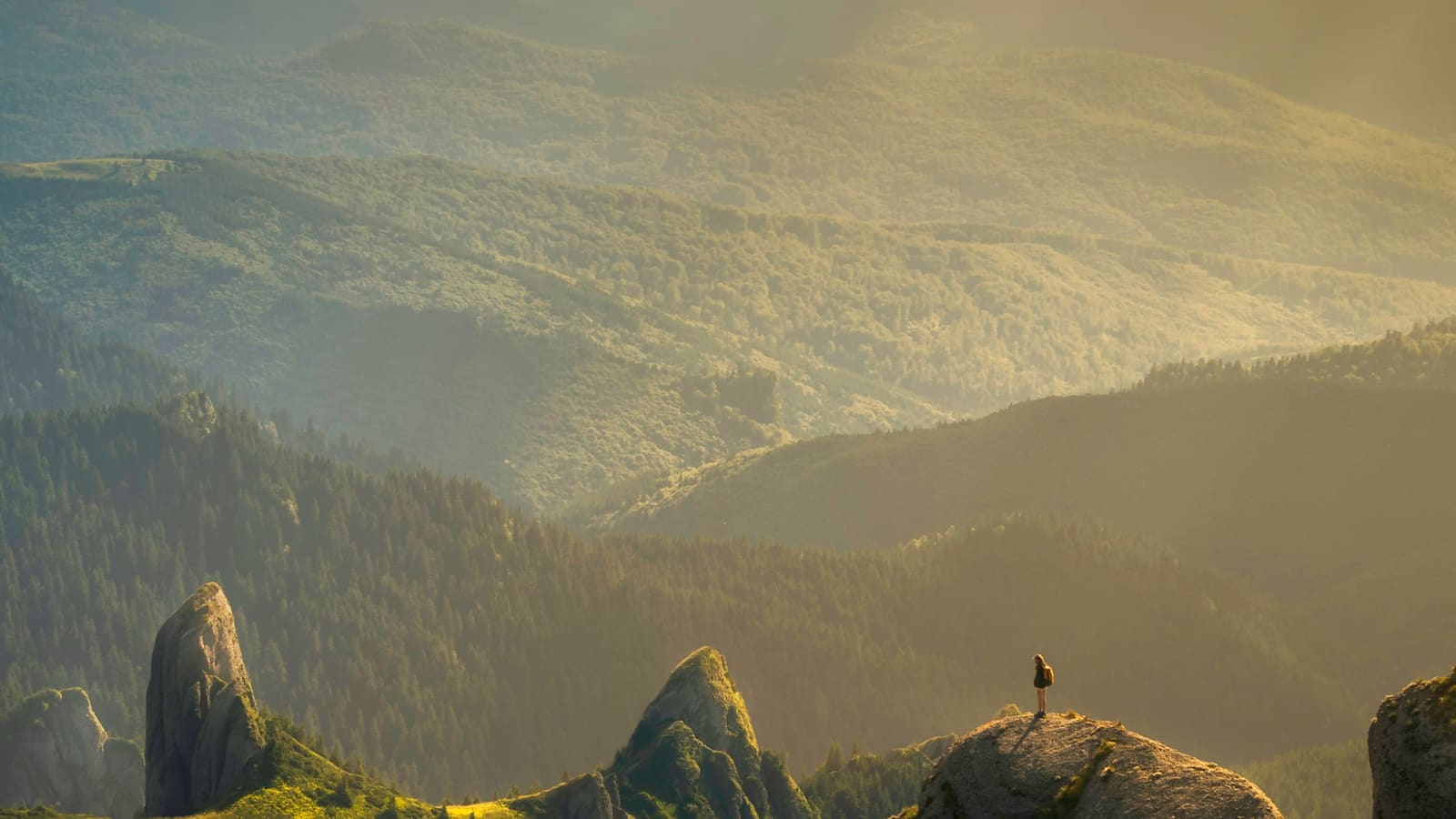 Golden evening light over rugged green cliffs with a lone hiker standing on a rock spire