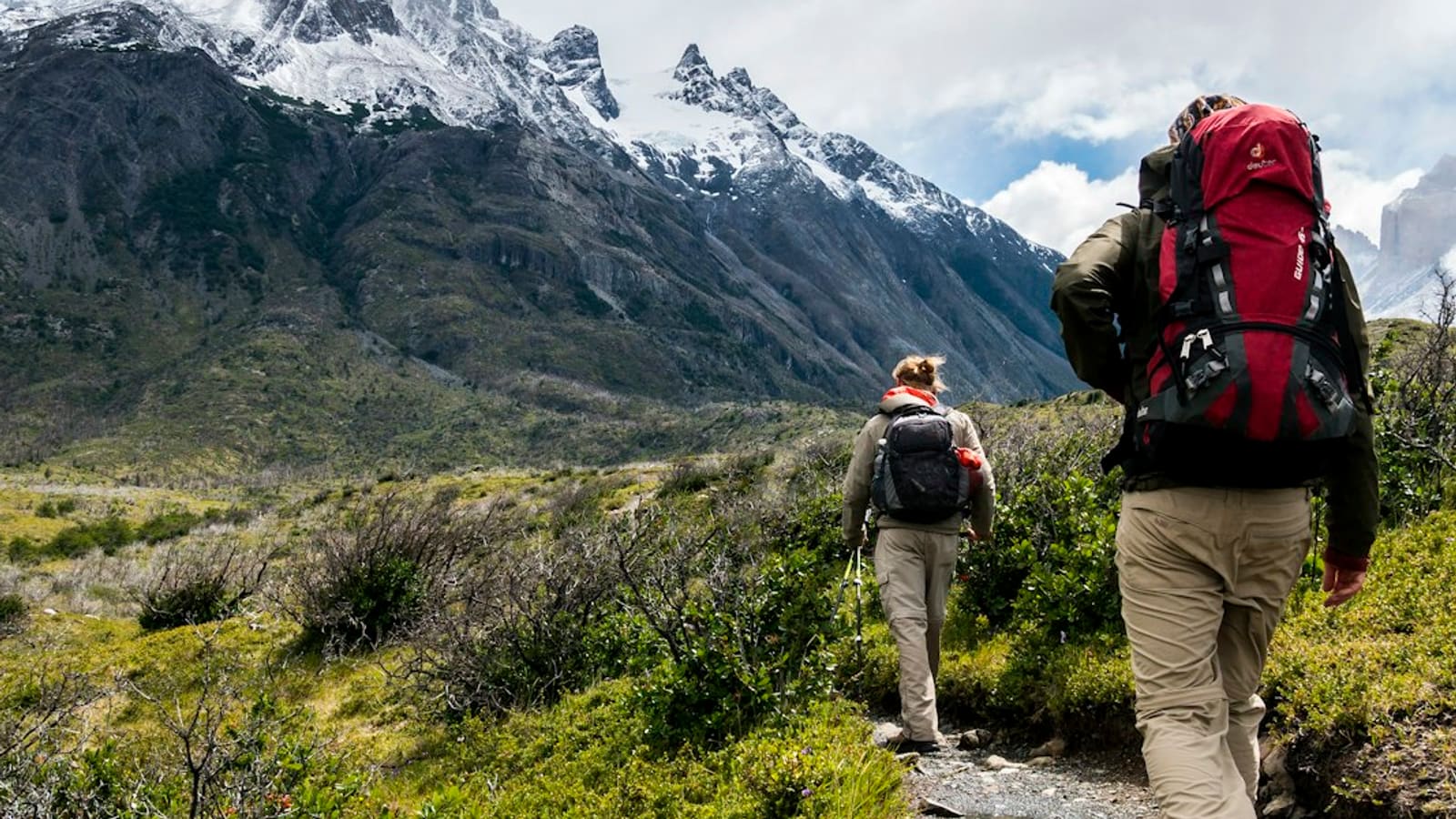 A backpacker with a large red pack hiking a trail toward a snow-capped peak