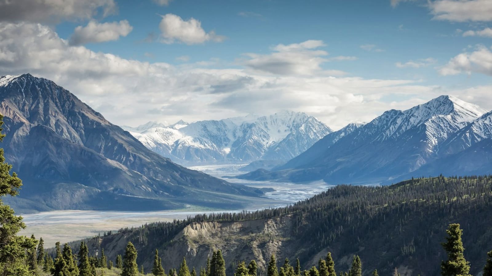 Wide mountain valley with a river, forest, and snowy summits under a bright sky