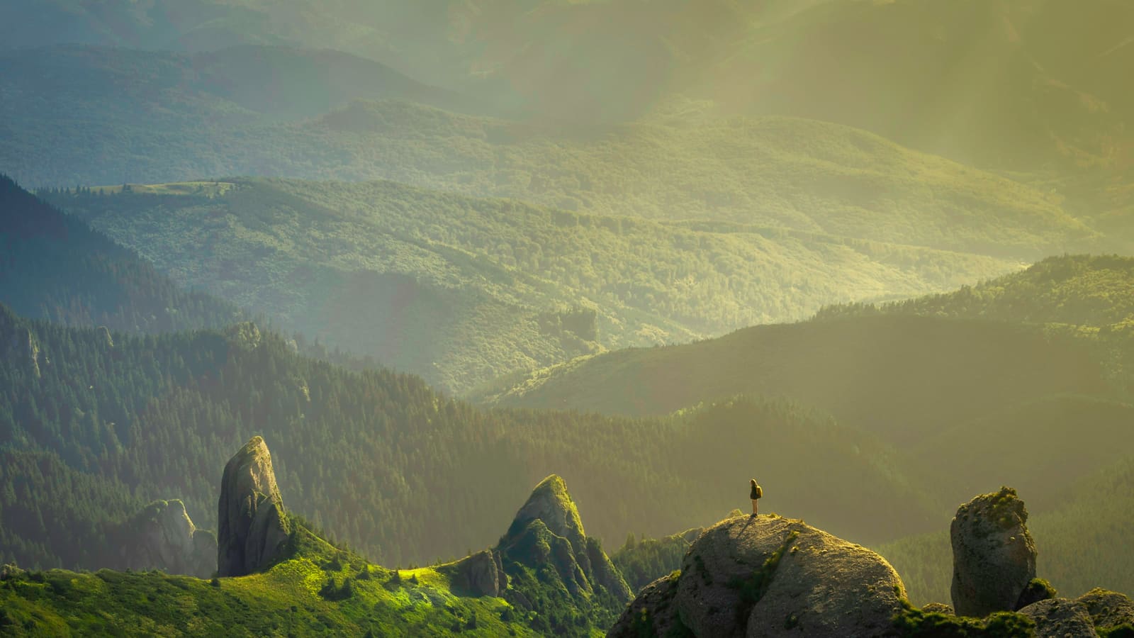 Golden evening light over rugged green cliffs with a lone hiker standing on a rock spire