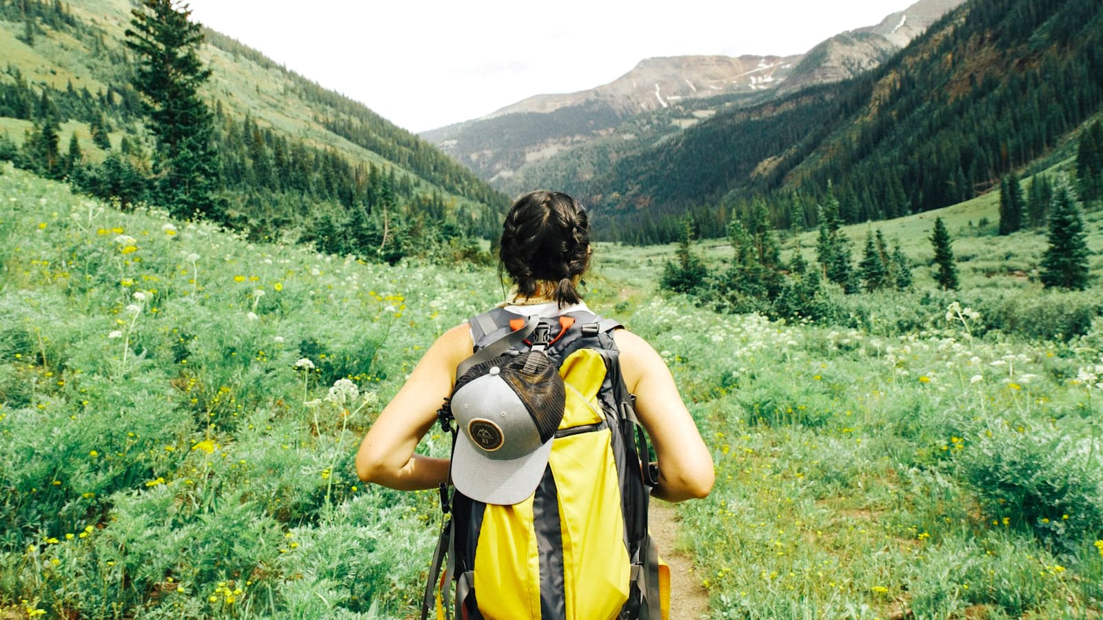 A backpacker with a yellow pack and sun hat looking across a green mountain valley