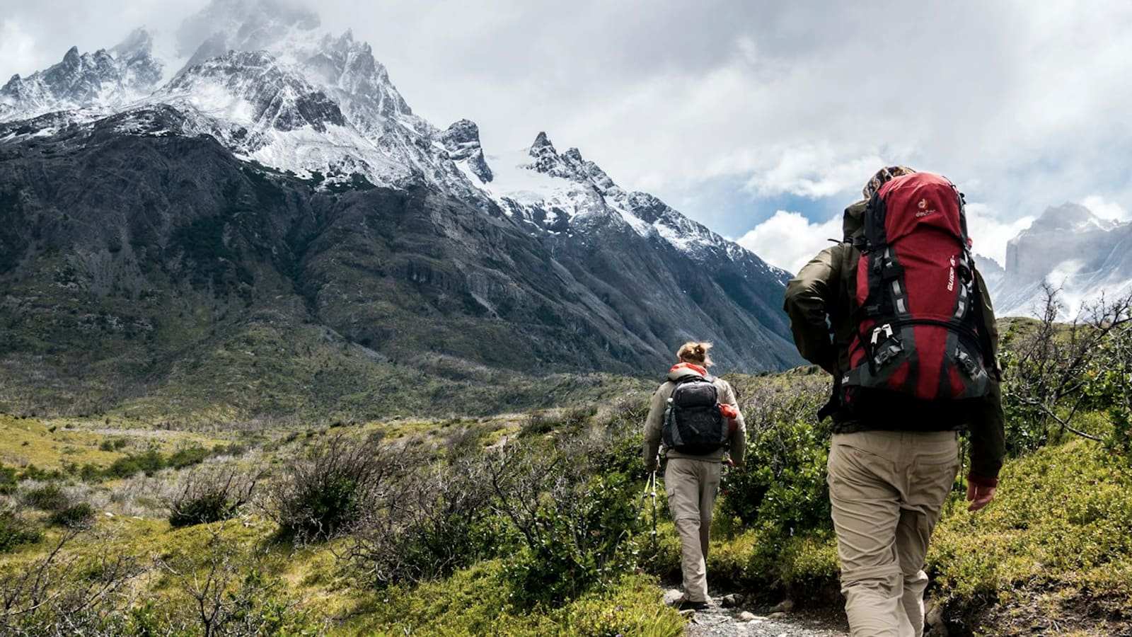 A backpacker with a large red pack hiking a trail toward a snow-capped peak