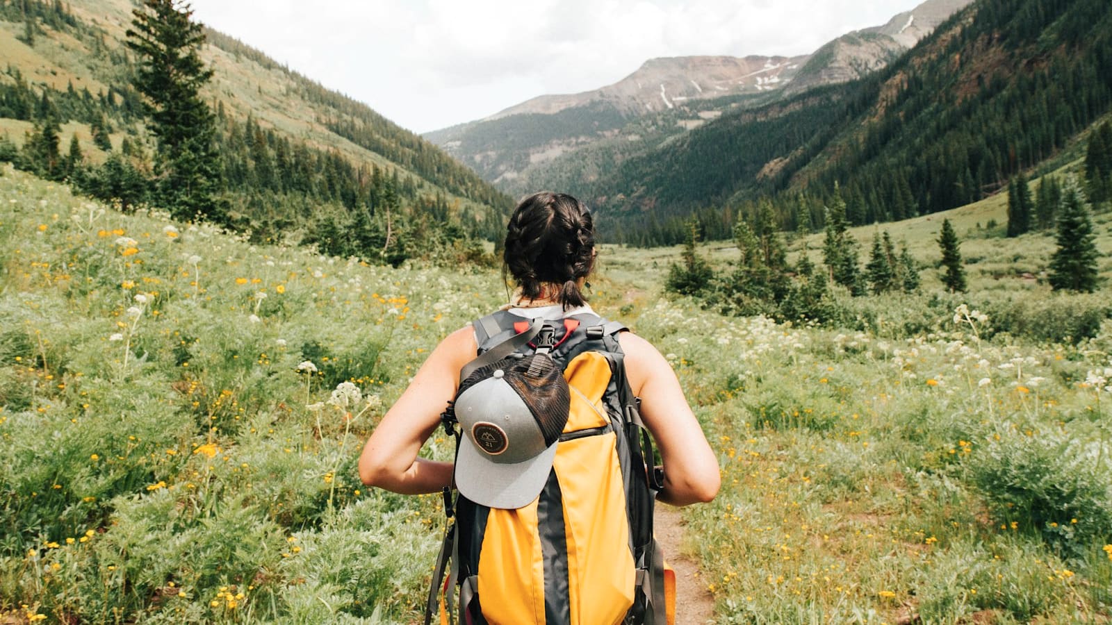 A backpacker with a yellow pack and sun hat looking across a green mountain valley