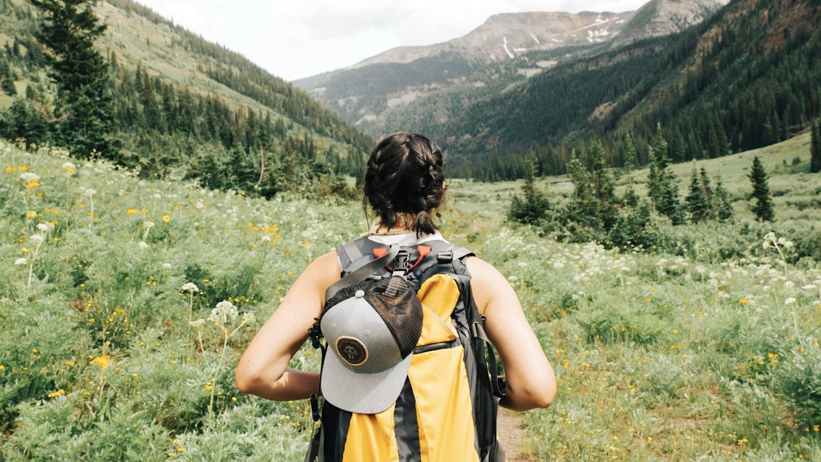 A backpacker with a yellow pack and sun hat looking across a green mountain valley