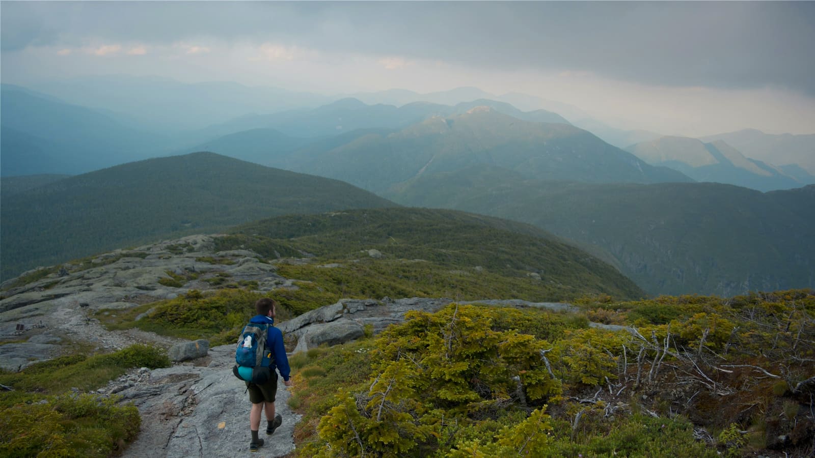 A hiker with a daypack climbing a rocky alpine trail above treeline in green mountains