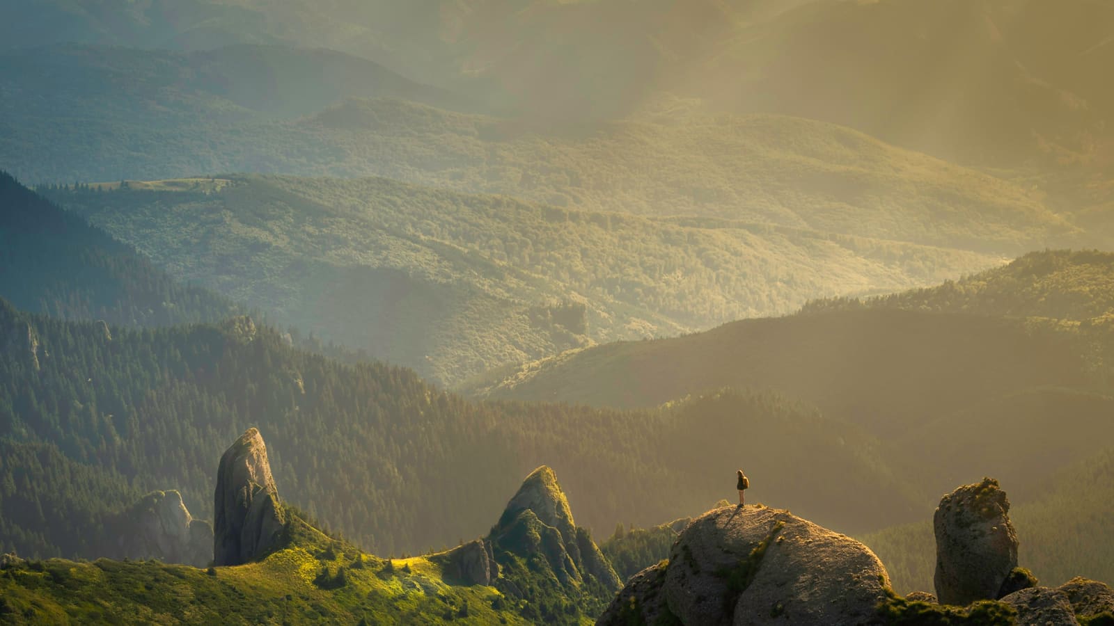 Golden evening light over rugged green cliffs with a lone hiker standing on a rock spire