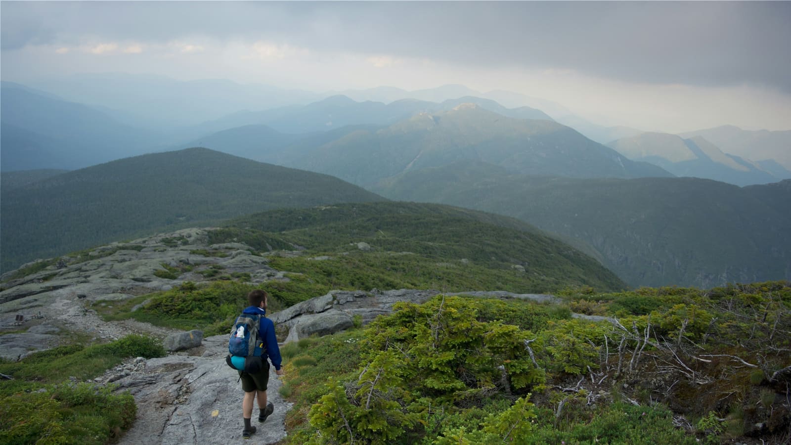 A hiker with a blue pack walking a rocky ridge trail through green mountains under a moody sky