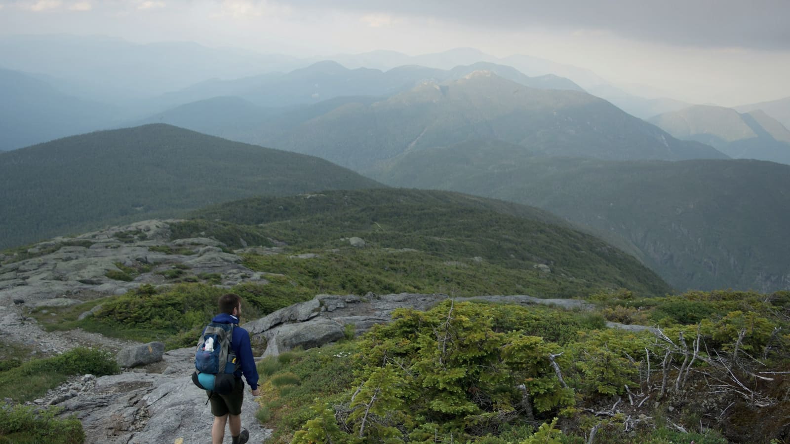 A hiker with a daypack climbing a rocky alpine trail above treeline in green mountains