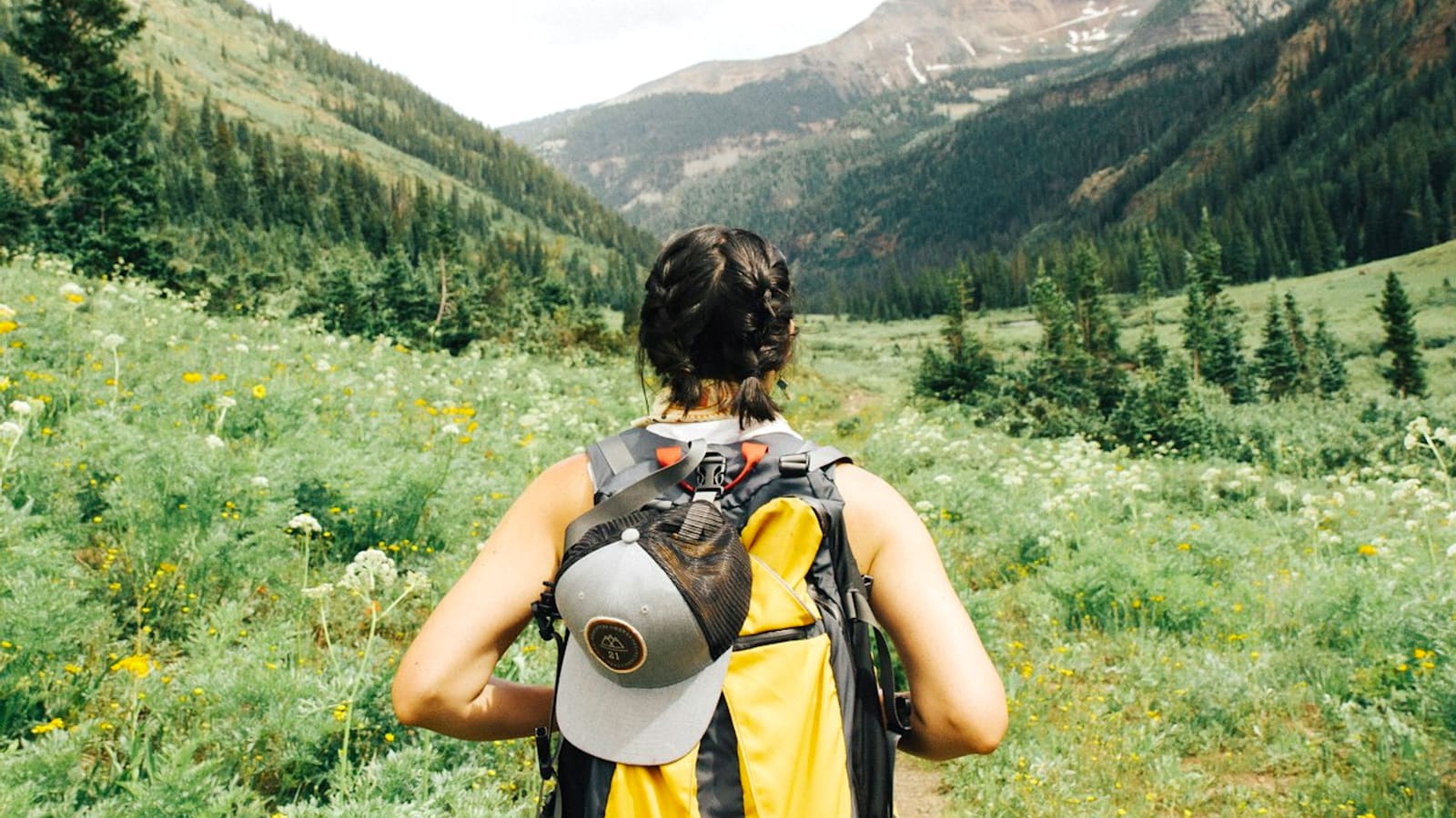 A backpacker with a yellow pack and sun hat looking across a green mountain valley